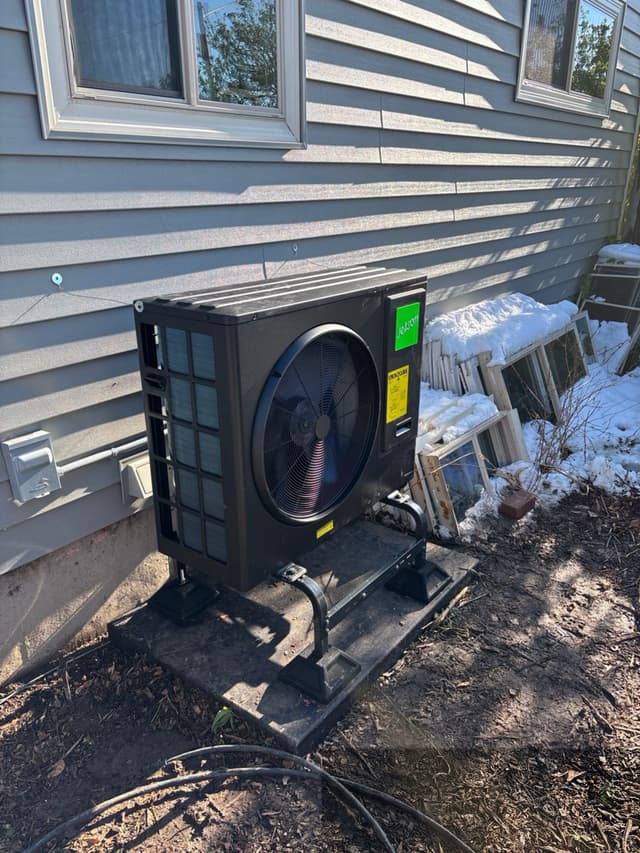 Black Jetson Air 3-ton heat pump condenser on a metal stand along the side of a gray house in Denver, CO, with an electrical disconnect box mounted to the wall nearby.
