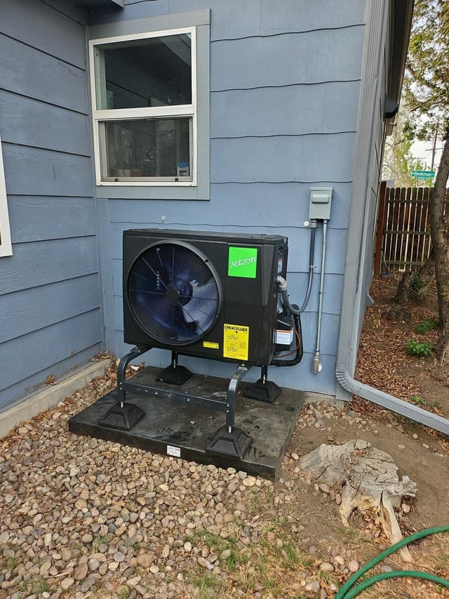A 2-ton Jetson Air central ducted heat pump mounted on a black metal stand on a concrete pad against the blue-gray lap siding of a Denver, CO home, with conduit and refrigerant lines running up the wall to a gray disconnect box.