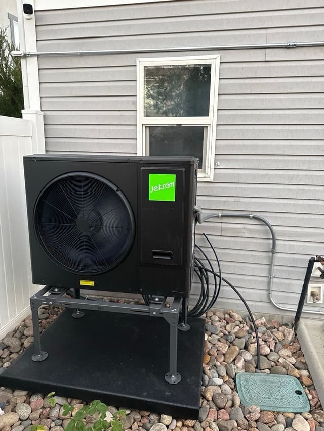 A black 3-ton Jetson Air central ducted heat pump mounted on an elevated stand against a gray-sided Denver home, with conduit running along the wall and a basement window visible above.