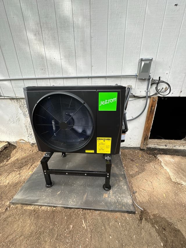 Jetson Air 2-ton heat pump condenser mounted against white vertical siding at a home in Westwood, Denver, with refrigerant lines running to a wall-mounted disconnect box.