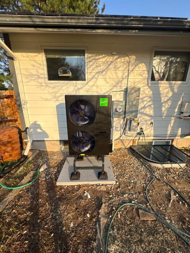 A black Jetson Air 5-ton heat pump on a concrete pad with leg risers against a cream-colored single-story home in Englewood, CO, with an electrical disconnect box and conduit runs on the wall to the right.