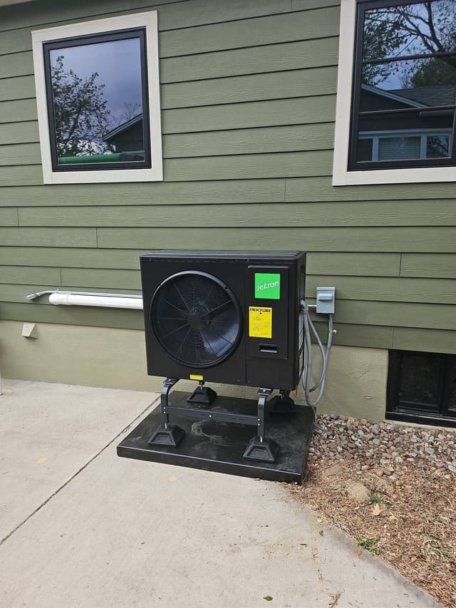 Jetson Air 3-ton heat pump condenser mounted on a black pad along the side of a green lap-sided house in Englewood, CO, with PVC line set cover and electrical conduit connected to a wall-mounted disconnect box.
