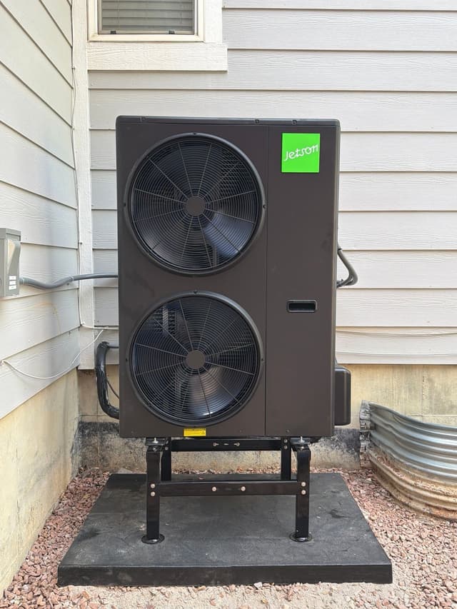 5-ton Jetson Air central ducted heat pump mounted on an elevated stand against a light gray lap-siding house in Erie, CO, with red gravel landscaping and neatly run refrigerant lines.