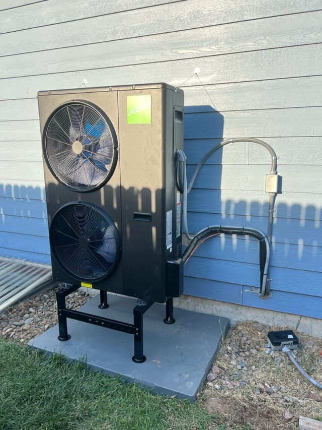 A black Jetson Air 4-ton heat pump mounted on a metal stand over a concrete pad against blue horizontal siding in Evans, CO, with refrigerant lines and conduit running neatly to a wall-mounted disconnect box.