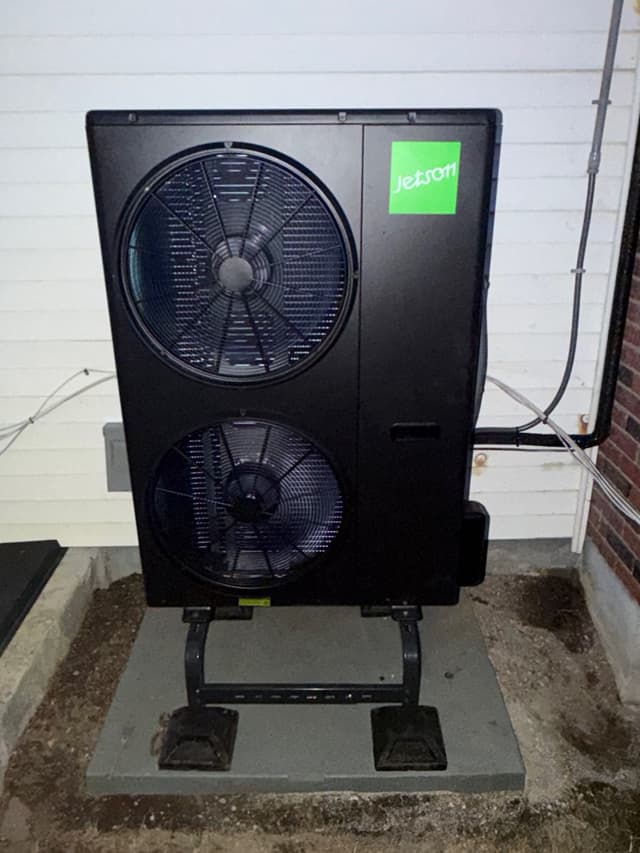 Jetson Air 4-ton dual-fan outdoor heat pump condenser mounted on a riser stand atop a concrete pad between a white-sided wall and a brick corner of a house in Everett, MA.