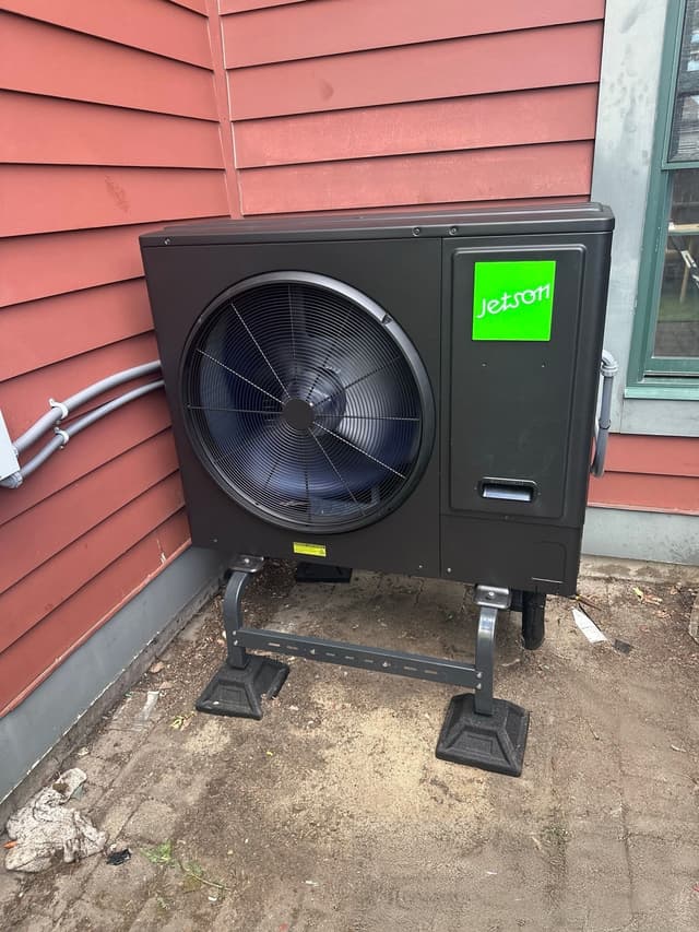 A black Jetson Air 3-ton central ducted heat pump installed against red horizontal siding near a green-trimmed window in Everett, MA.