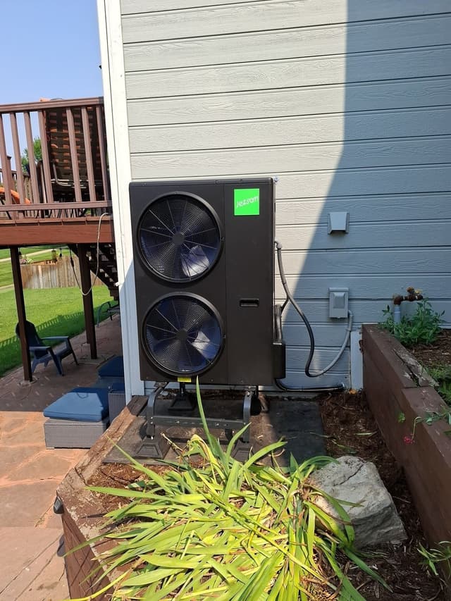 Black Jetson Air 4-ton heat pump installed against light-blue vinyl siding at a Fort Collins home, with flexible conduit running to an electrical disconnect box and a raised garden bed with ornamental grasses in the foreground.