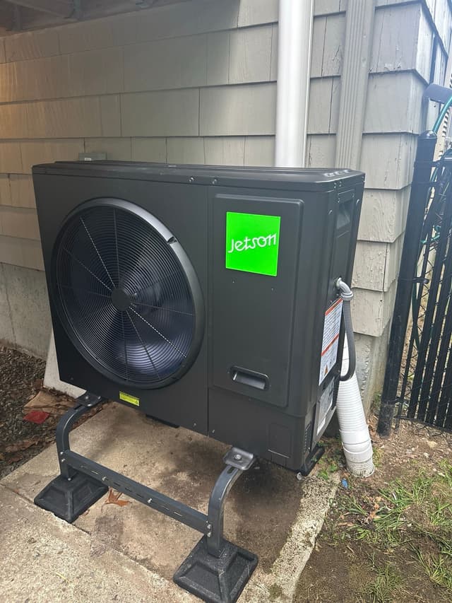 A black Jetson Air 3-ton central ducted heat pump mounted outside a gray-sided home in Foxborough, MA, with a black iron fence visible to the right.