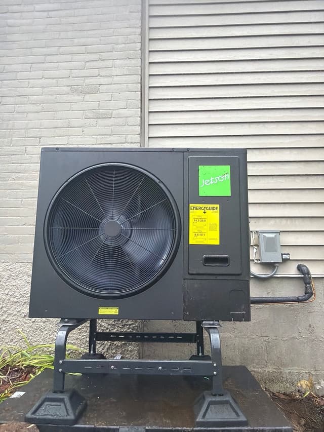 Black Jetson Air 3-ton central ducted heat pump outdoor unit installed against a grey vinyl and brick exterior wall in Gardner, MA, with refrigerant lines and disconnect box visible.