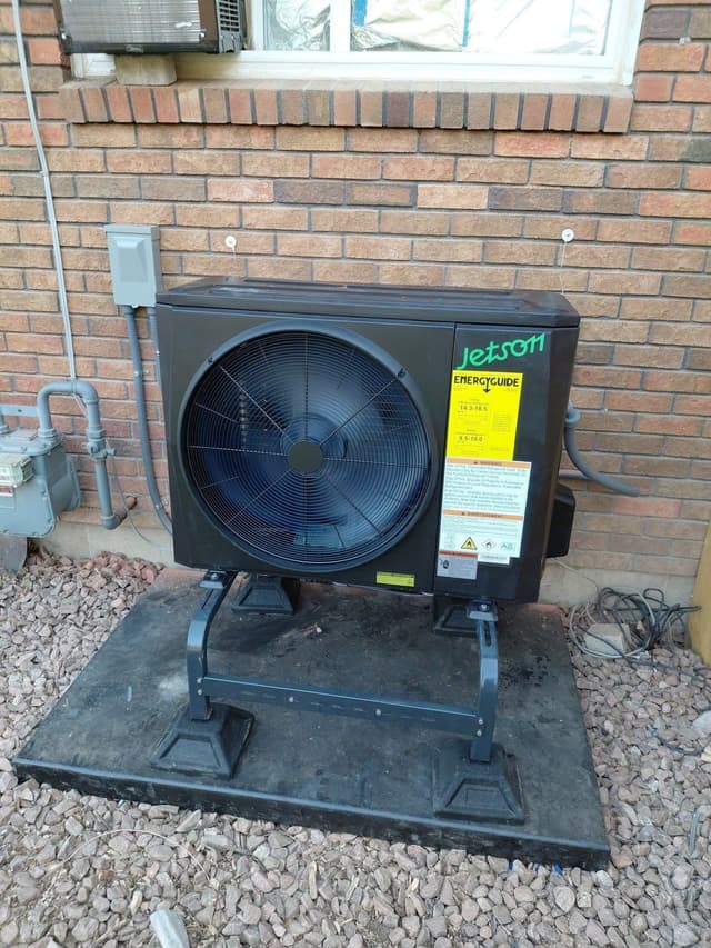 Jetson Air 2-ton central ducted heat pump installed against a red brick exterior wall in Greeley, with disconnect box and refrigerant lines visible.