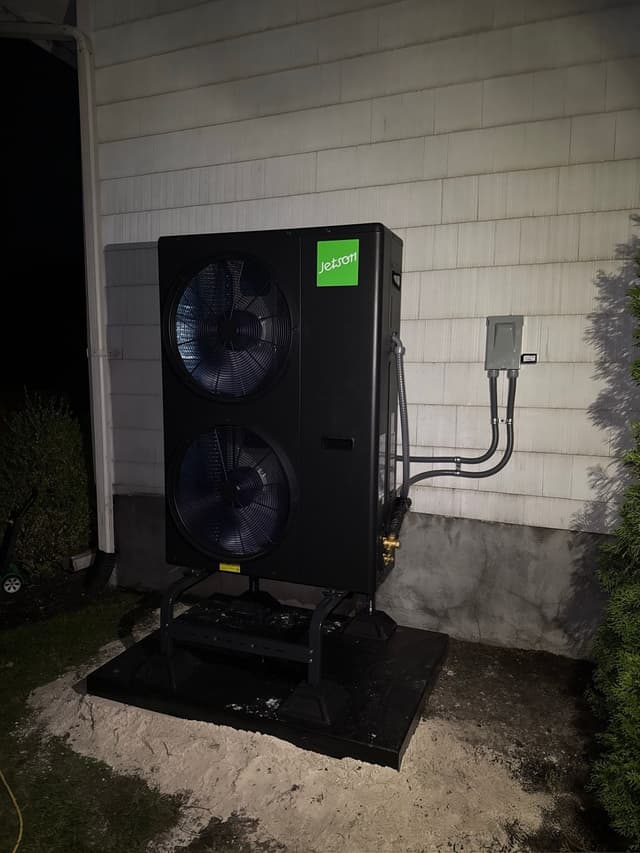 Large black Jetson Air 5-ton heat pump unit mounted against white shingled siding at a Harrison home, with two visible fan faces, the Jetson logo lit in green, and conduit running to a wall-mounted disconnect box.