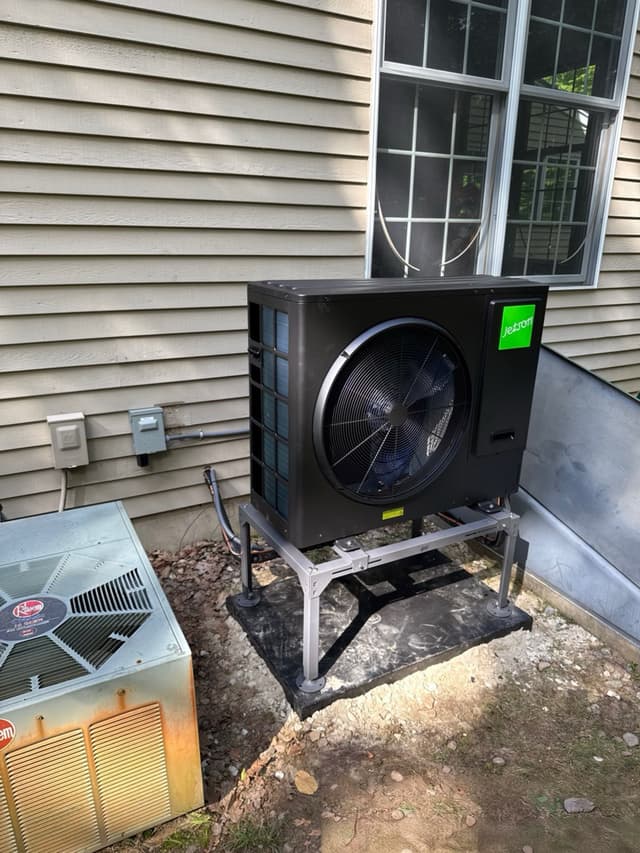 Black Jetson Air 3-ton heat pump installed beside the foundation of a gray-sided house in Harvard, MA, with electrical disconnects mounted on the siding and an old Rheem condenser visible nearby.