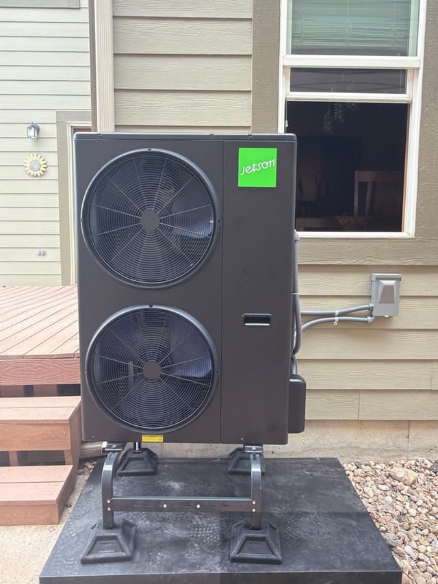 A black Jetson Air 4-ton central ducted heat pump installed against the tan lap siding of a Highlands Ranch home, with condenser lines and a disconnect box visible along the wall.