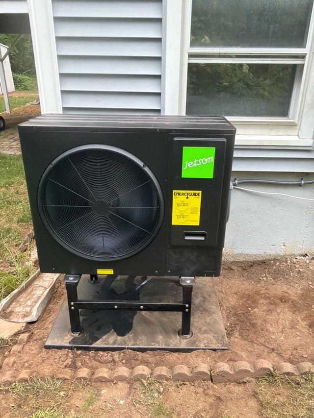 Black Jetson Air 3-ton heat pump mounted on an elevated stand over pavers against gray house siding in Hopedale, MA, with conduit lines running along the wall.