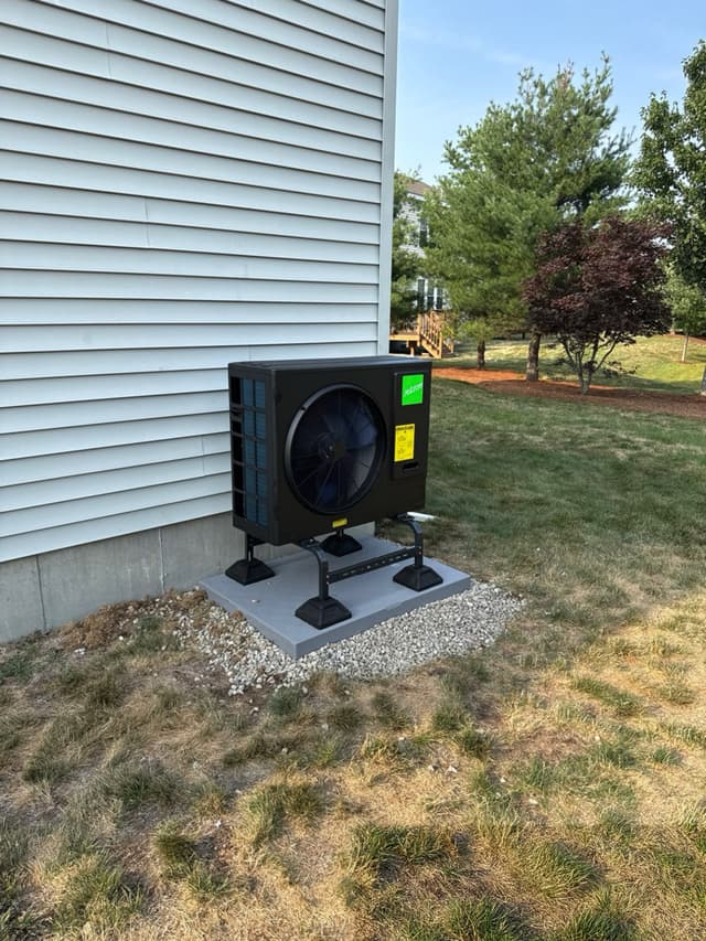 A black Jetson Air 3-ton central ducted heat pump condenser installed against light-gray vinyl siding at a home in Hopkinton, MA, with a well-kept lawn and mature trees in the background.