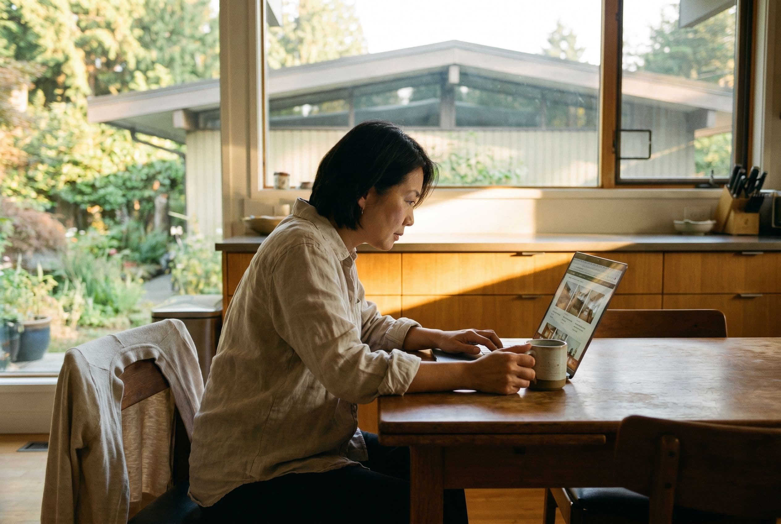 A homeowner comparing heat pump options on a laptop in a sunlit mid-century kitchen in Metro Vancouver