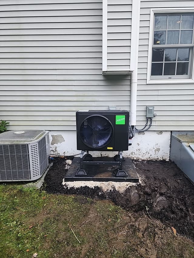 Black Jetson Air 3-ton central ducted heat pump installed on a concrete pad against gray vinyl siding in Katonah, NY, with electrical disconnect box and conduit mounted on the wall.