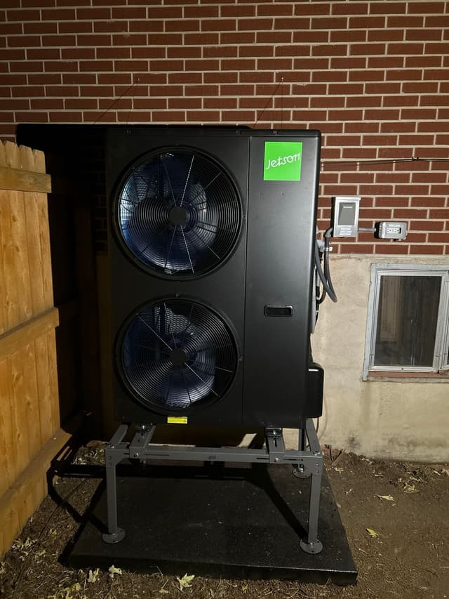 4-ton Jetson Air central ducted heat pump installed against a red brick foundation wall beside a wooden fence in Lakewood, with electrical disconnect and control panel mounted to the wall.