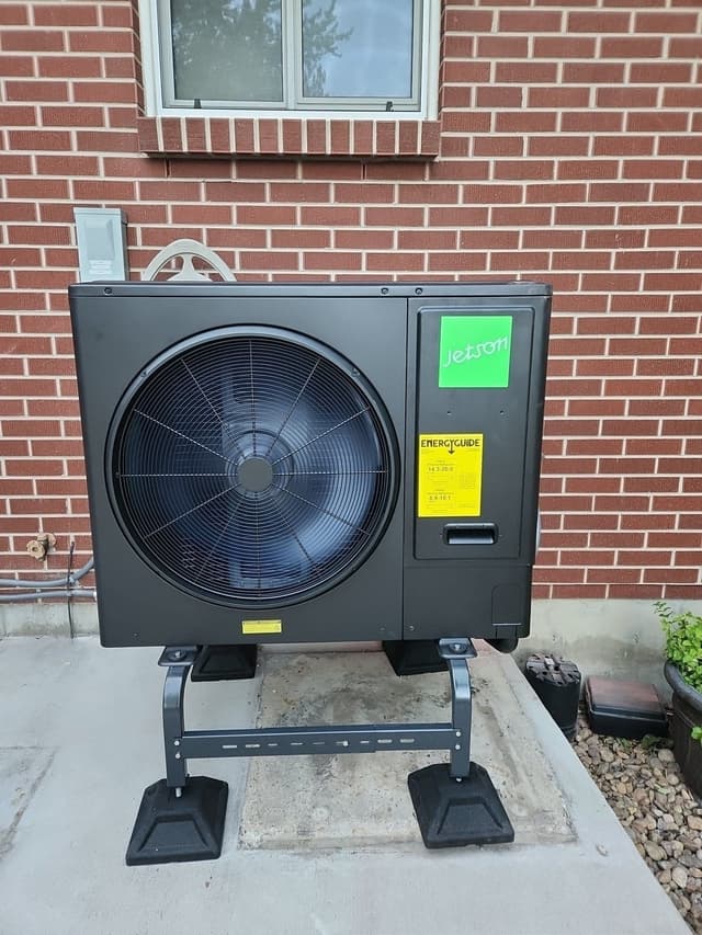 Jetson Air 3-ton heat pump outdoor unit mounted against a red brick wall in Lakewood, CO, with an electrical disconnect box visible to the left.