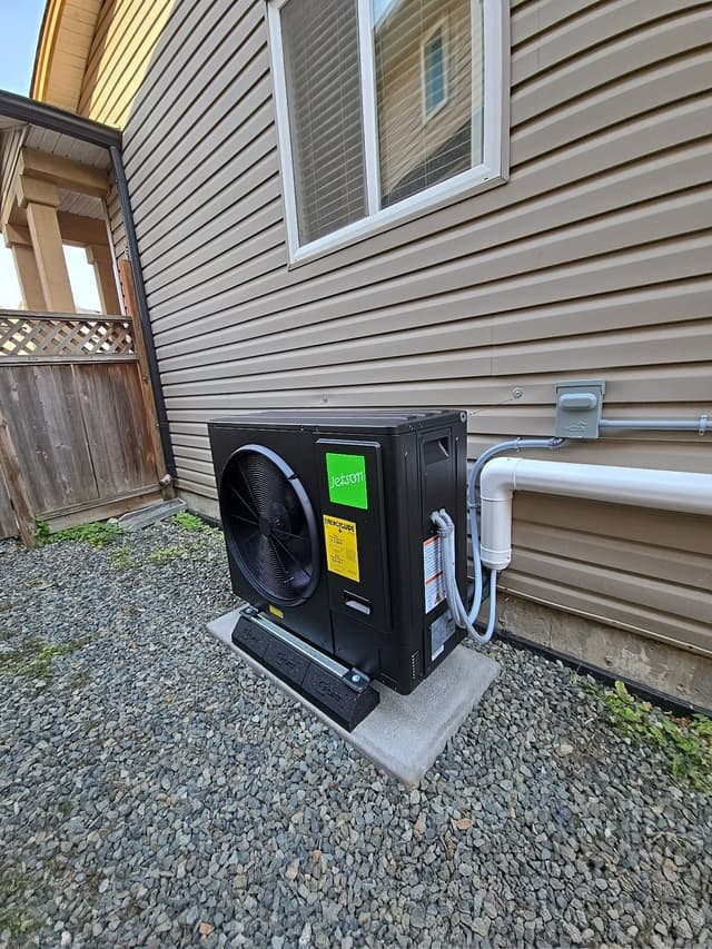 Black Jetson Air 3-ton heat pump condenser installed on a concrete pad in a gravel side yard against tan lap siding, with refrigerant lines and conduit running up to a wall-mounted disconnect box in Langley.