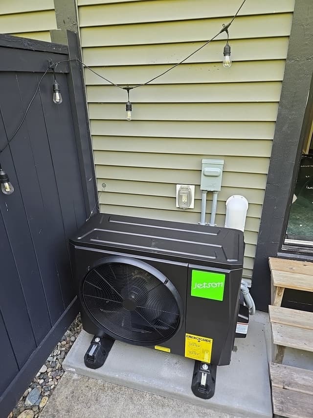 A black Jetson Air 2-ton heat pump condenser on a concrete pad in a corner between a dark fence and green-sided house wall, with electrical conduit, a disconnect box, and string lights above, in Walnut Grove, Langley Township, BC.