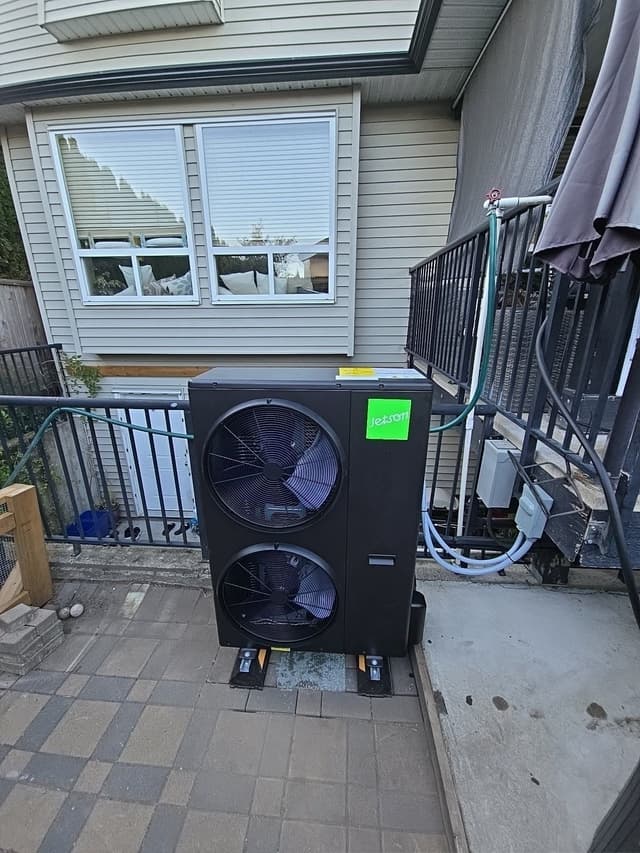 A black Jetson Air dual-fan 4-ton heat pump mounted on anti-vibration feet on a paved backyard patio, with refrigerant lines and conduit running to a nearby disconnect box, at a home in Aldergrove, Langley Township, BC.