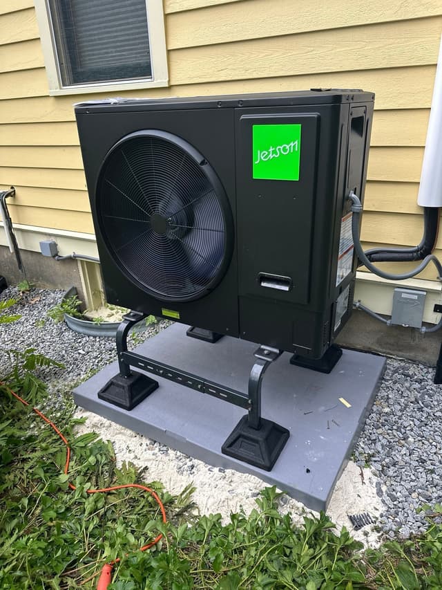 Black Jetson Air 3-ton heat pump condenser installed along the side of a yellow-sided house in Lexington, MA, with refrigerant lines running neatly toward the home.