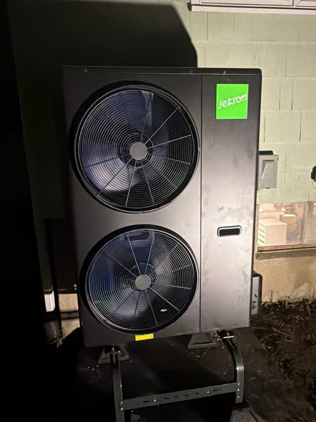 Dual-fan black Jetson Air 4-ton heat pump condenser installed against a concrete block wall in Lexington, MA, photographed at night with the green Jetson logo visible on the unit.