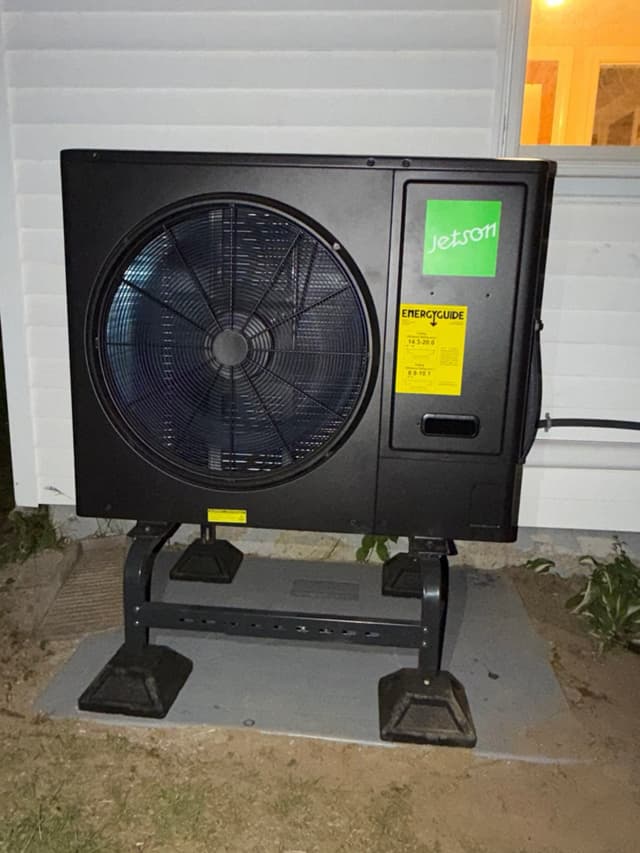 Jetson Air 3-ton central ducted heat pump unit mounted on a metal riser beside white house siding, photographed straight-on at night in Lexington, MA.