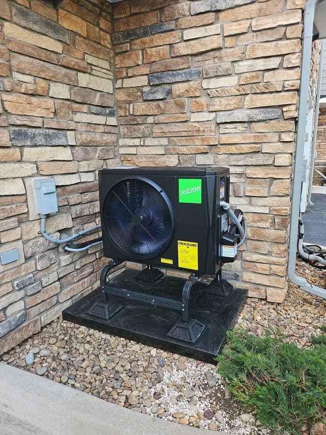 2-ton Jetson Air heat pump condenser on a metal stand in a gravel bed against the stone-veneer exterior corner of a home in Littleton, CO, with refrigerant lines and electrical disconnect visible.
