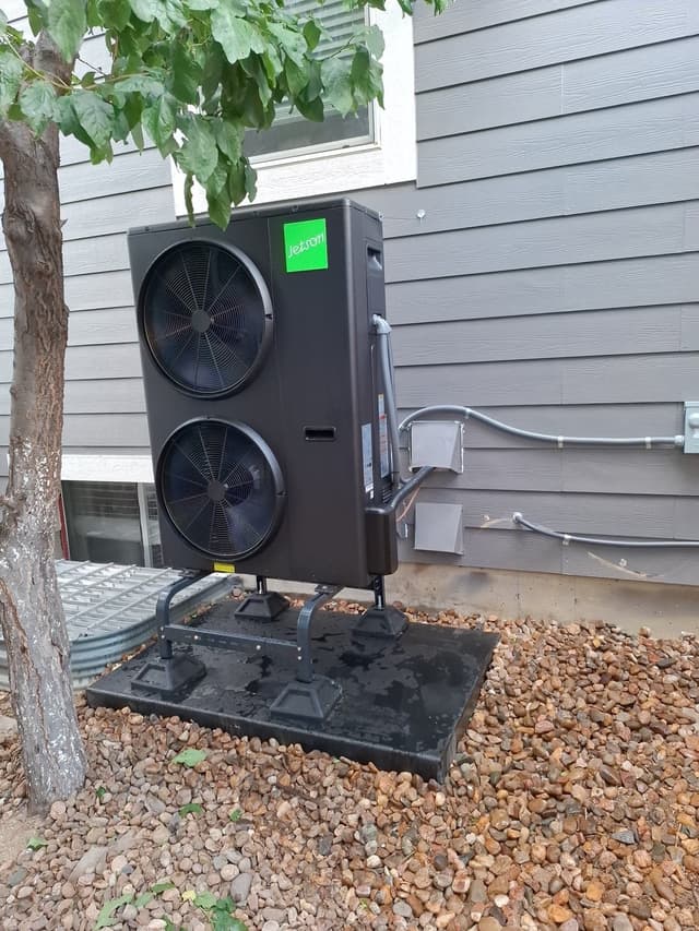 Jetson Air 3-ton heat pump on an elevated stand in a gravel bed beside a gray-sided house in Littleton, CO, with conduit and disconnect box visible on the wall.