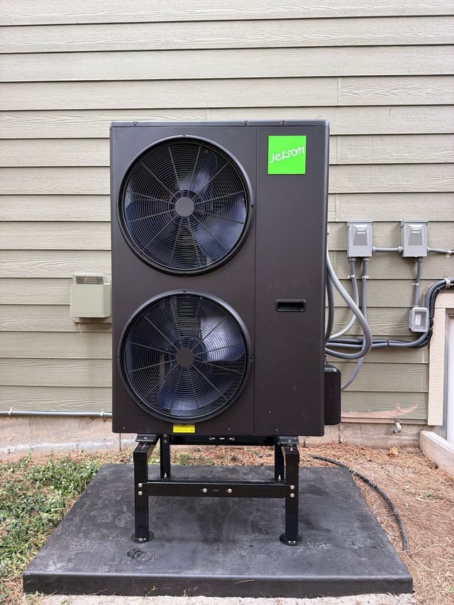 Jetson Air 4-ton central ducted heat pump mounted on a steel stand against light-grey horizontal siding at a home in Littleton, MA, with electrical disconnect boxes and conduit visible on the wall beside it.