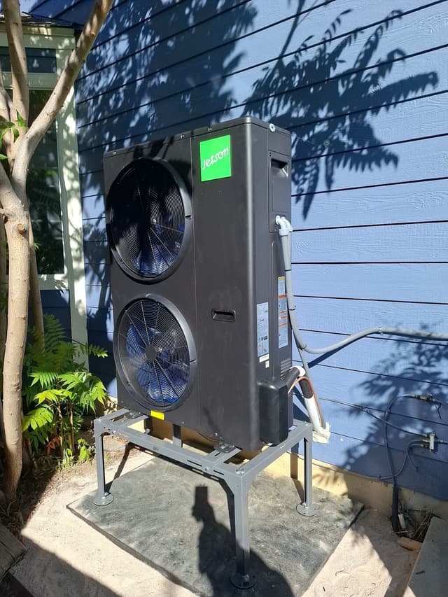Black Jetson Air central ducted heat pump condenser mounted on a steel stand against blue horizontal siding at a home in Littleton, MA.