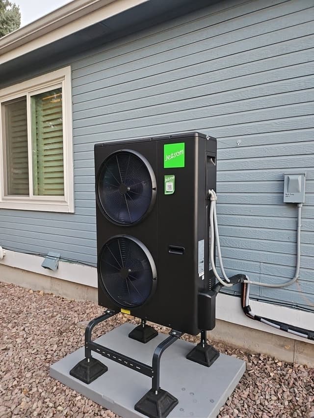 Black Jetson Air 4-ton heat pump mounted on a metal stand over a concrete pad on gravel, with conduit and refrigerant lines running to a disconnect box, against a light-blue sided house in Littleton, CO.