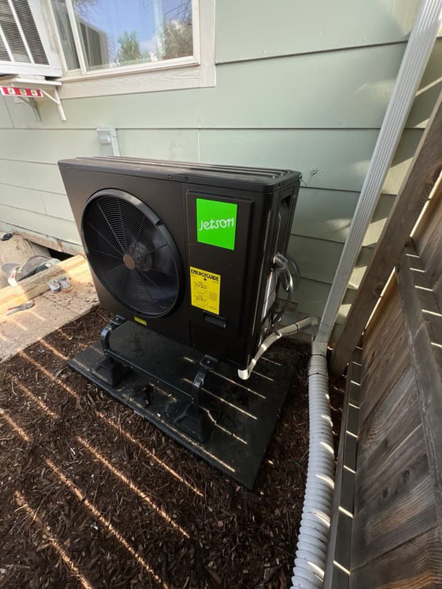 Jetson Air 3-ton heat pump mounted on a black equipment stand in a wood-chip mulch bed along the side of a light green lap-siding house in Longmont, CO, with refrigerant lines and conduit running down the right side of the unit.