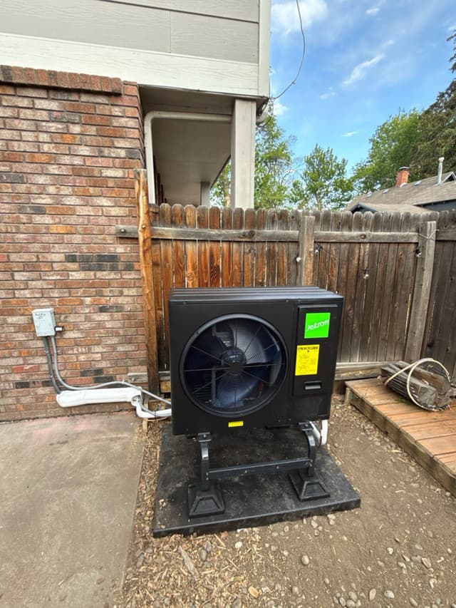 Black Jetson Air 3-ton outdoor heat pump unit on a pad beside a brick house in Longmont, CO, with electrical conduit running to a disconnect box on the wall and a wooden privacy fence in the background.
