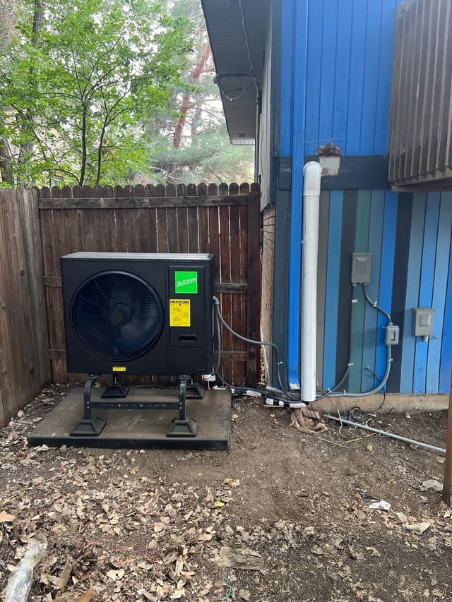 A black Jetson Air 3-ton heat pump condenser installed in a side yard in Longmont, Colorado, with refrigerant lines and electrical conduit running along a blue vertical-board building wall.