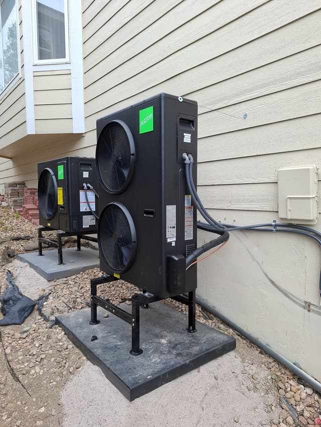 Two black Jetson Air heat pump condensers installed side by side along the exterior wall of a tan lap siding house in Louisville, CO, with refrigerant lines and conduit running neatly to an electrical box.