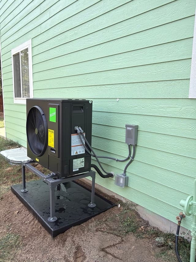 Black Jetson Air 3-ton heat pump mounted on an elevated stand against light-green lap siding of a home in Loveland, with conduit and wiring running neatly to a disconnect box on the house wall.