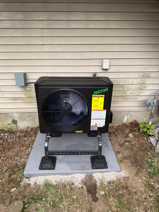 Jetson Air 2-ton central ducted heat pump mounted against beige lap siding on a completed install in Lowell, MA, with disconnect box and gas pipe visible to either side.