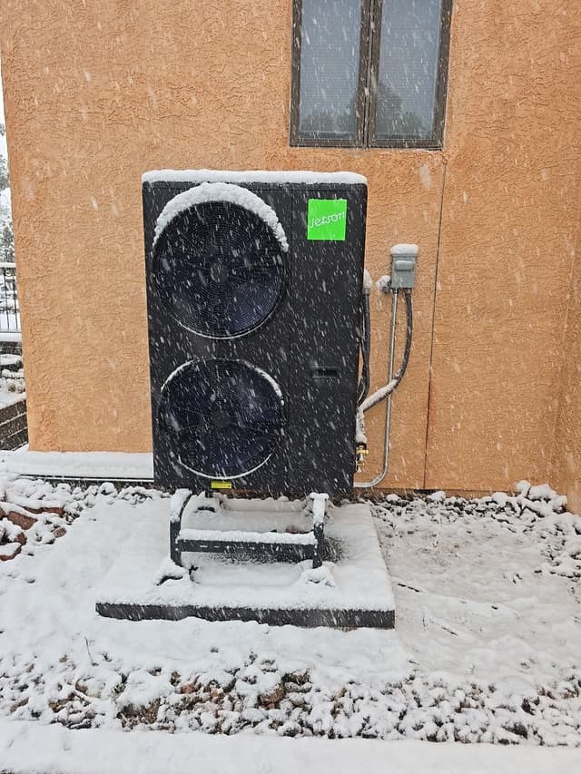 A Jetson Air 5-ton heat pump on a raised metal pad in front of a tan stucco wall in Manitou Springs, CO, with snow falling heavily and accumulating on top of the unit and on the ground.