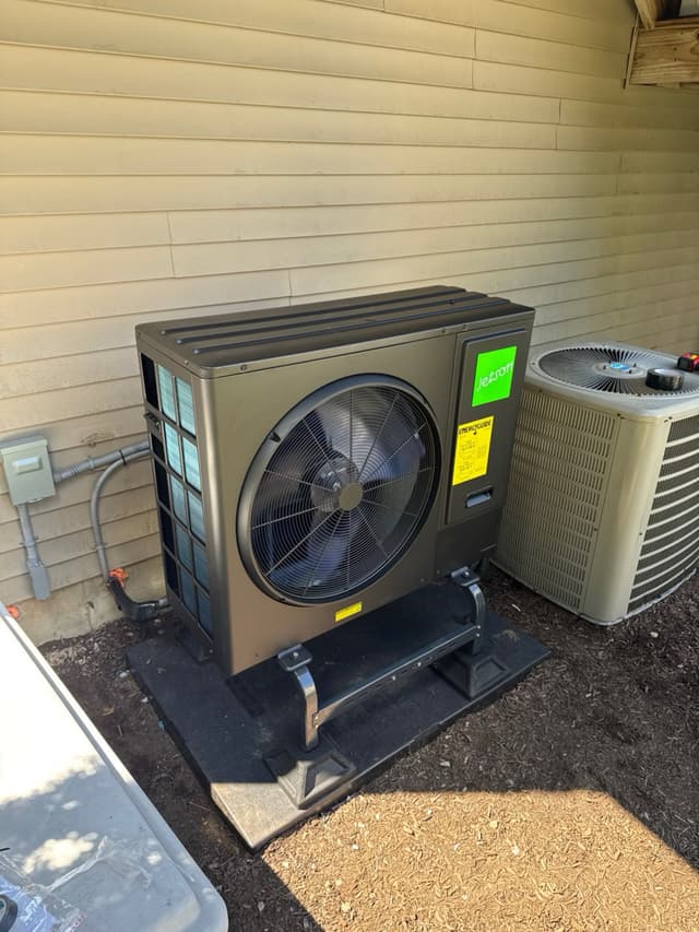 A charcoal Jetson Air 3-ton central ducted heat pump on a plastic pad beside a vinyl-sided house in Mansfield, MA, with electrical conduit running to a disconnect box and an existing AC condenser visible to the right.