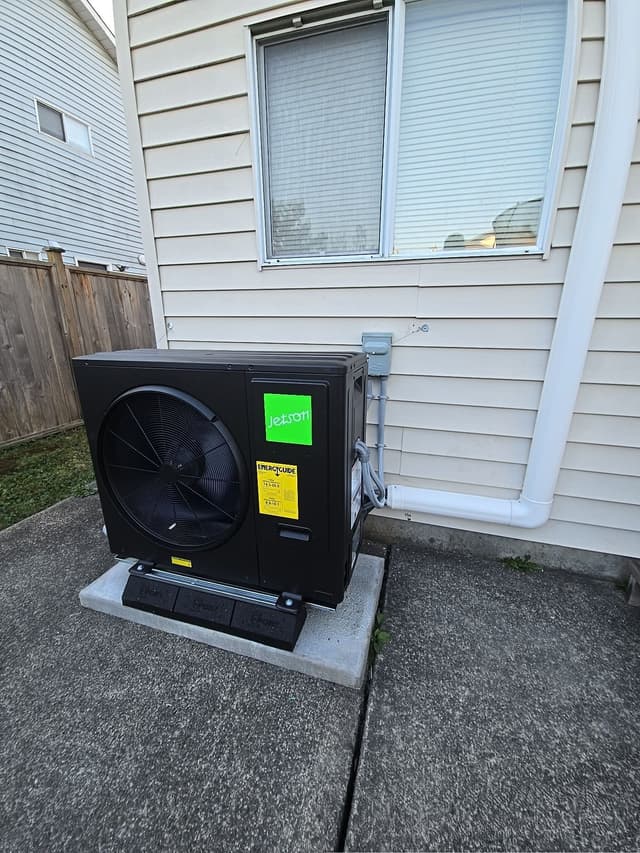 Black Jetson Air 3-ton central ducted heat pump installed against beige vinyl siding on a home in Port Hammond, Maple Ridge, with electrical conduit and disconnect box neatly run up the wall.