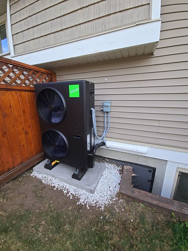 A black Jetson Air 4-ton heat pump unit installed against beige vinyl siding beside a wood-lattice fence in Albion, Maple Ridge, with conduit and piping running neatly to a wall-mounted disconnect box.