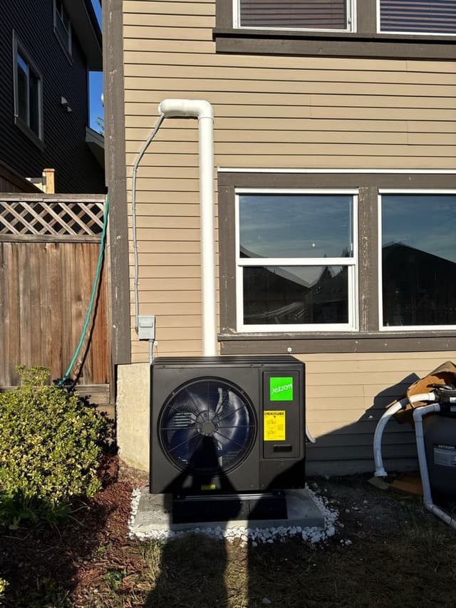 A black Jetson Air 3-ton central ducted heat pump installed on a white gravel pad along the side of a tan two storey home in Maple Ridge, BC, with white PVC conduit running up the wall and pool equipment partially visible to the right.