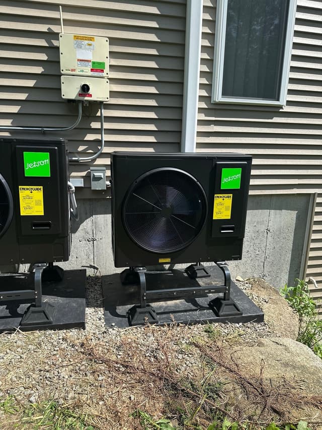 Two black Jetson Air outdoor heat pump condenser units installed side by side against a gray-sided house foundation in Milford, with solar disconnect hardware mounted on the siding above.