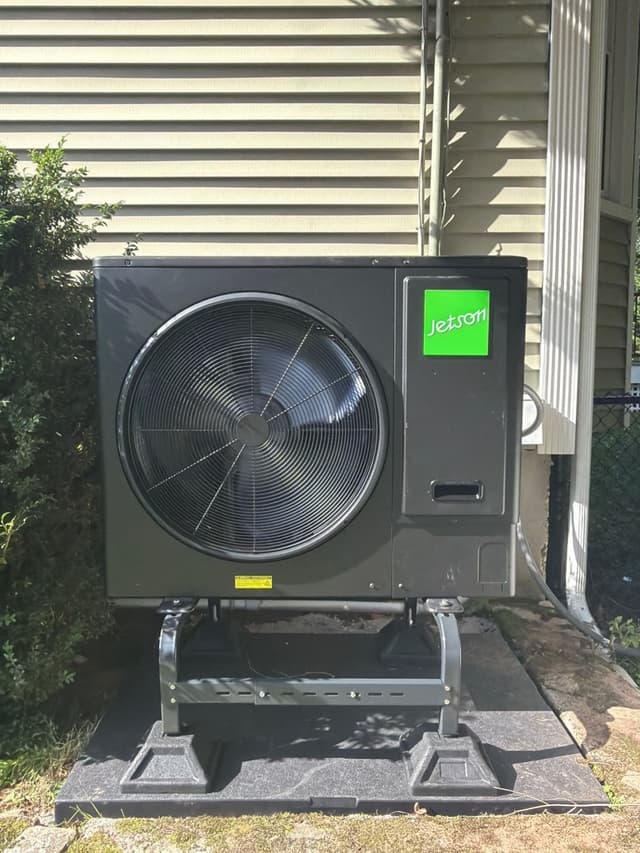 3-ton Jetson Air central ducted heat pump mounted on a black metal riser stand on a concrete surface beside a tan vinyl-sided house in Natick, MA.