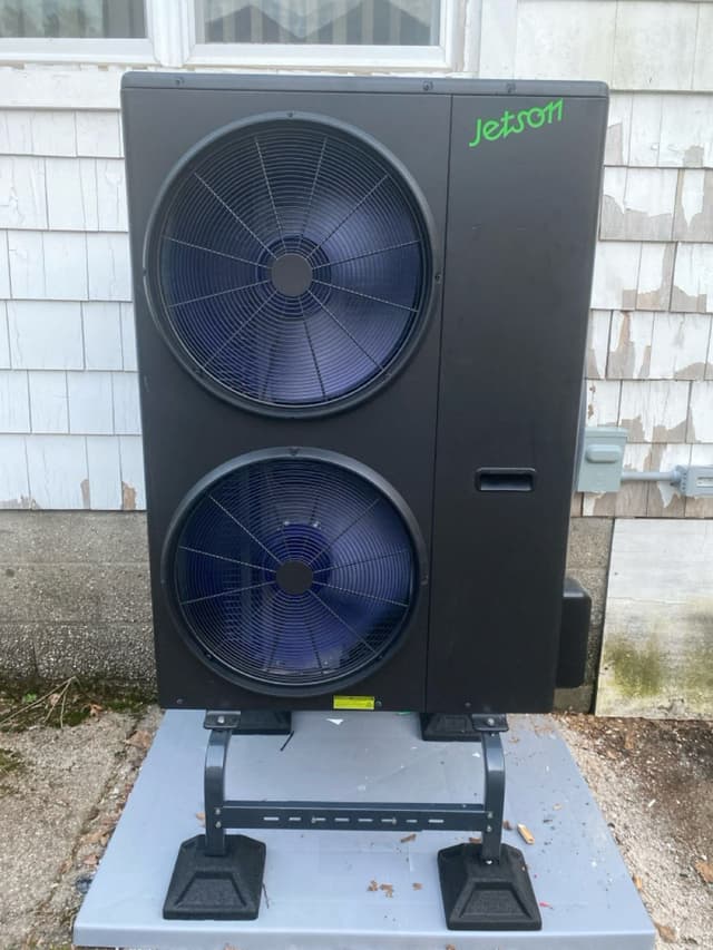 Jetson Air 4-ton heat pump mounted on a pad and riser against the gray-shingle siding of a home in Newton, MA, with an electrical panel visible to the right.