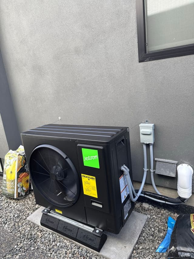 Black Jetson Air 3-ton central ducted heat pump installed against a grey stucco exterior wall in Lynn Valley, North Vancouver, with line-set piping running to a disconnect box mounted on the wall.