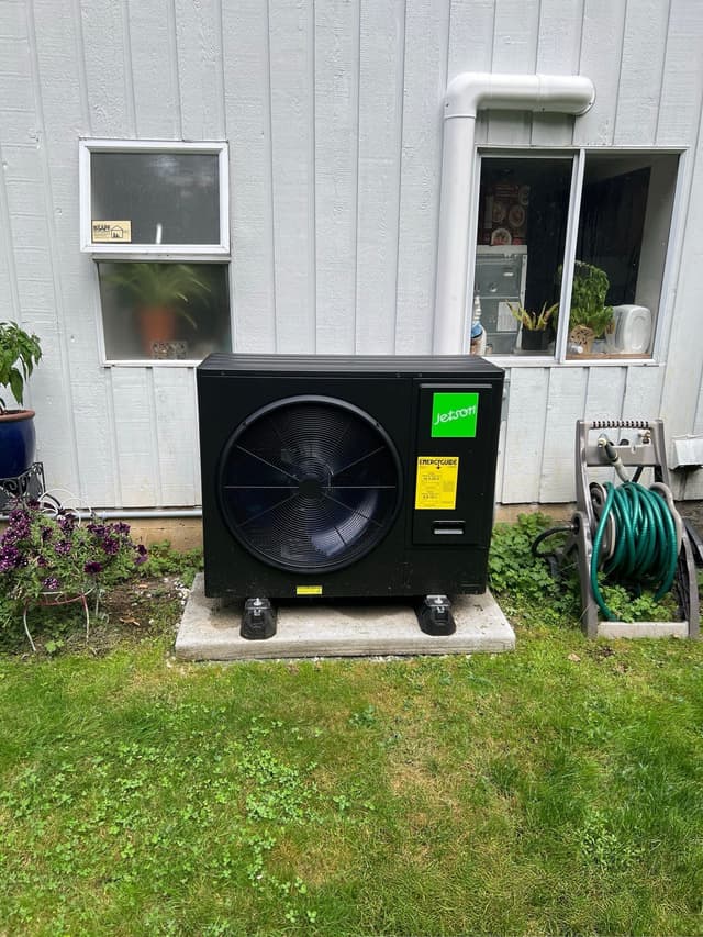 Black Jetson Air 3-ton central ducted heat pump installed on a concrete pad beside a white-sided house in Lynn Valley, North Vancouver, with flowering plants visible to the left.